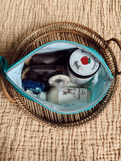 The inside view of a medium andiamo bag to demonstrate how much it can hold. It has baby shoes, a fig bar snack, a pacifier, along with a pump bottle of french diapering cream and a container of lotion. The bag is on top of a woven basket, on top of a beige blanket.