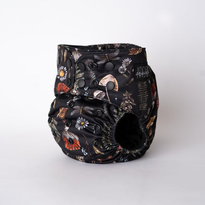 A pocket style cloth diaper from Arcadia’s Nursery shown at a slight front angle in the Midnight Flora print. The deep black background is scattered with detailed florals, mushrooms, moths, and foliage in muted tones of rust, ivory, sage, and gold. Adjustable snap closures and softly gathered leg elastics highlight the contoured fit and dark, botanical aesthetic.