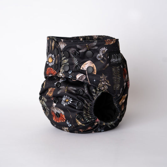 A pocket style cloth diaper from Arcadia’s Nursery shown at a slight front angle in the Midnight Flora print. The deep black background is scattered with detailed florals, mushrooms, moths, and foliage in muted tones of rust, ivory, sage, and gold. Adjustable snap closures and softly gathered leg elastics highlight the contoured fit and dark, botanical aesthetic.