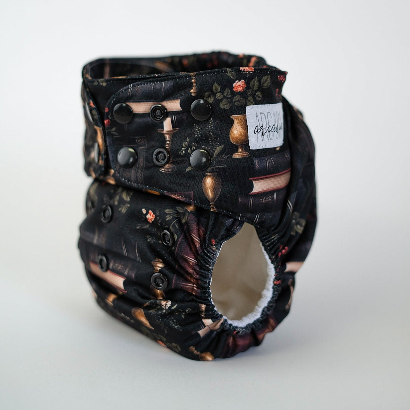 A pocket diaper with a dark pattern featuring books and academic elements, with a brand label showing the name 'Arcadia'.