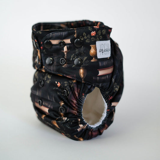 A pocket diaper with a dark pattern featuring books and academic elements, with a brand label showing the name 'Arcadia'.