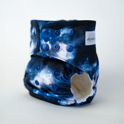 A pocket style cloth diaper from Arcadia’s Nursery shown at a slight front angle in the Milky Way print. The cosmic fabric displays rich blues, inky shadows, and scattered star like details for a bold celestial look. Coordinating snap closures along the waist and rise provide an adjustable fit, and the curved leg openings highlight the sculpted, performance focused design.