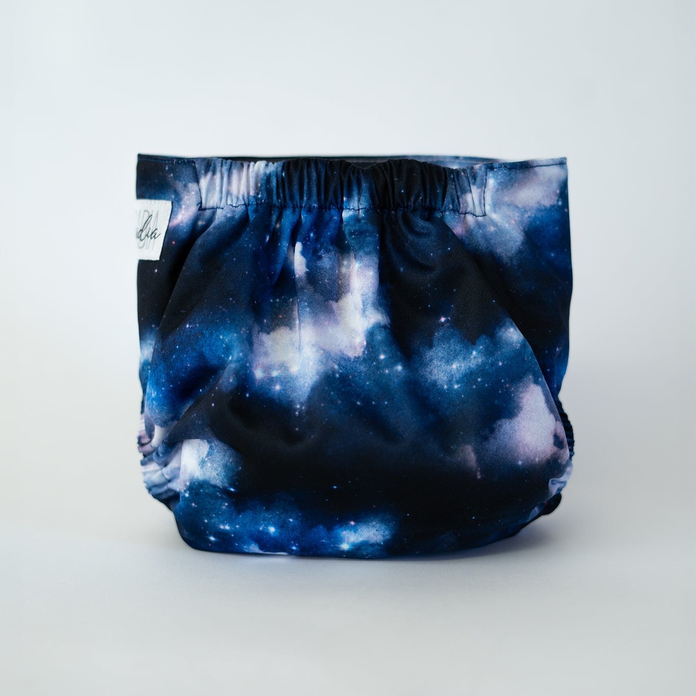 The back view of an Arcadia’s Nursery pocket cloth diaper in the Milky Way print, displayed against a white background. The fabric features a dramatic cosmic design with deep navy and midnight blue tones blended with cloudy white and soft star speckling. Gathered elastic at the waist and legs creates a secure, contoured fit while showcasing the swirling galaxy inspired artwork.