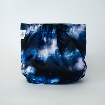 The back view of an Arcadia’s Nursery pocket cloth diaper in the Milky Way print, displayed against a white background. The fabric features a dramatic cosmic design with deep navy and midnight blue tones blended with cloudy white and soft star speckling. Gathered elastic at the waist and legs creates a secure, contoured fit while showcasing the swirling galaxy inspired artwork.