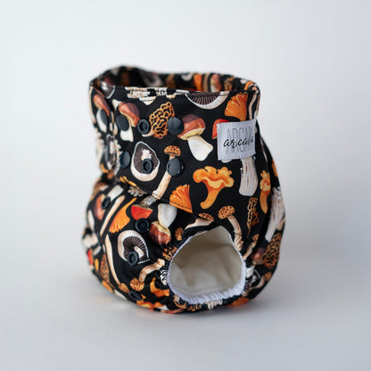 A cloth pocket diaper with a black background featuring a colorful mushroom print in earthy tones. It has black snaps for adjustable sizing, elasticized leg openings, and a soft interior. The Arcadia's Nursery logo is stitched on the front. Front angle view.