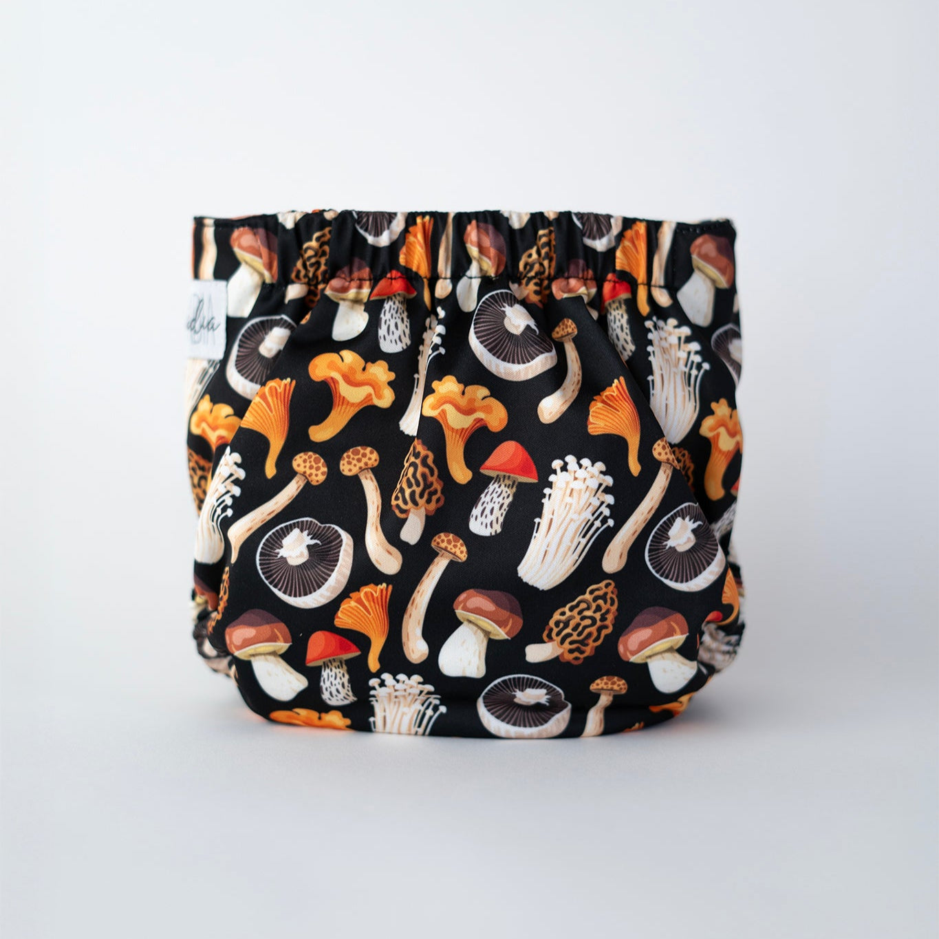 A cloth pocket diaper featuring a black background with a colorful mushroom print in earthy tones, including orange, brown, and beige. The design showcases various mushroom types in detail. The diaper has elastic along the back for a secure fit and the Arcadia's Nursery logo tag on the side. Rear view.