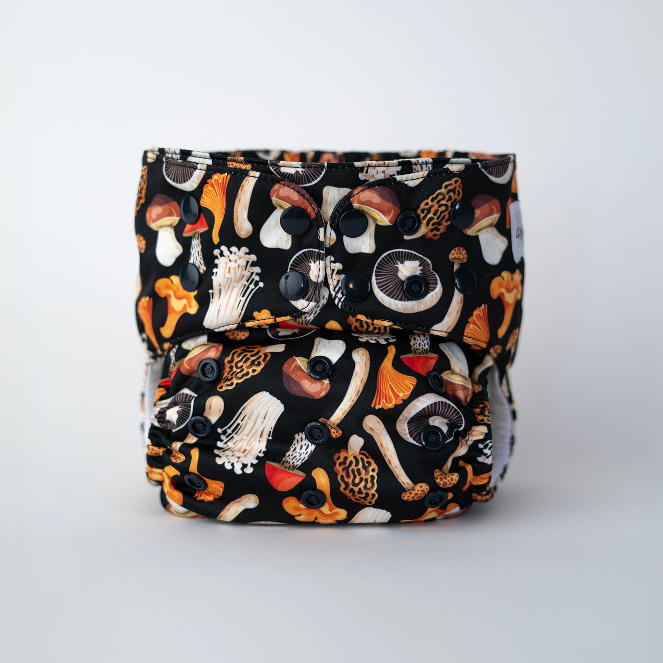 A cloth pocket diaper with a black background featuring a colorful mushroom print in earthy tones. It has black snaps for adjustable sizing, elasticized leg openings, and a soft interior. The Arcadia's Nursery logo is stitched on the front. Front VIew.