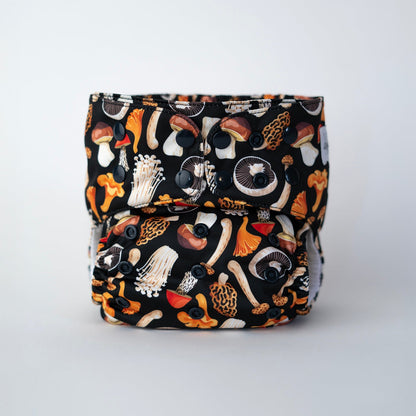 A cloth pocket diaper with a black background featuring a colorful mushroom print in earthy tones. It has black snaps for adjustable sizing, elasticized leg openings, and a soft interior. The Arcadia's Nursery logo is stitched on the front. Front VIew.
