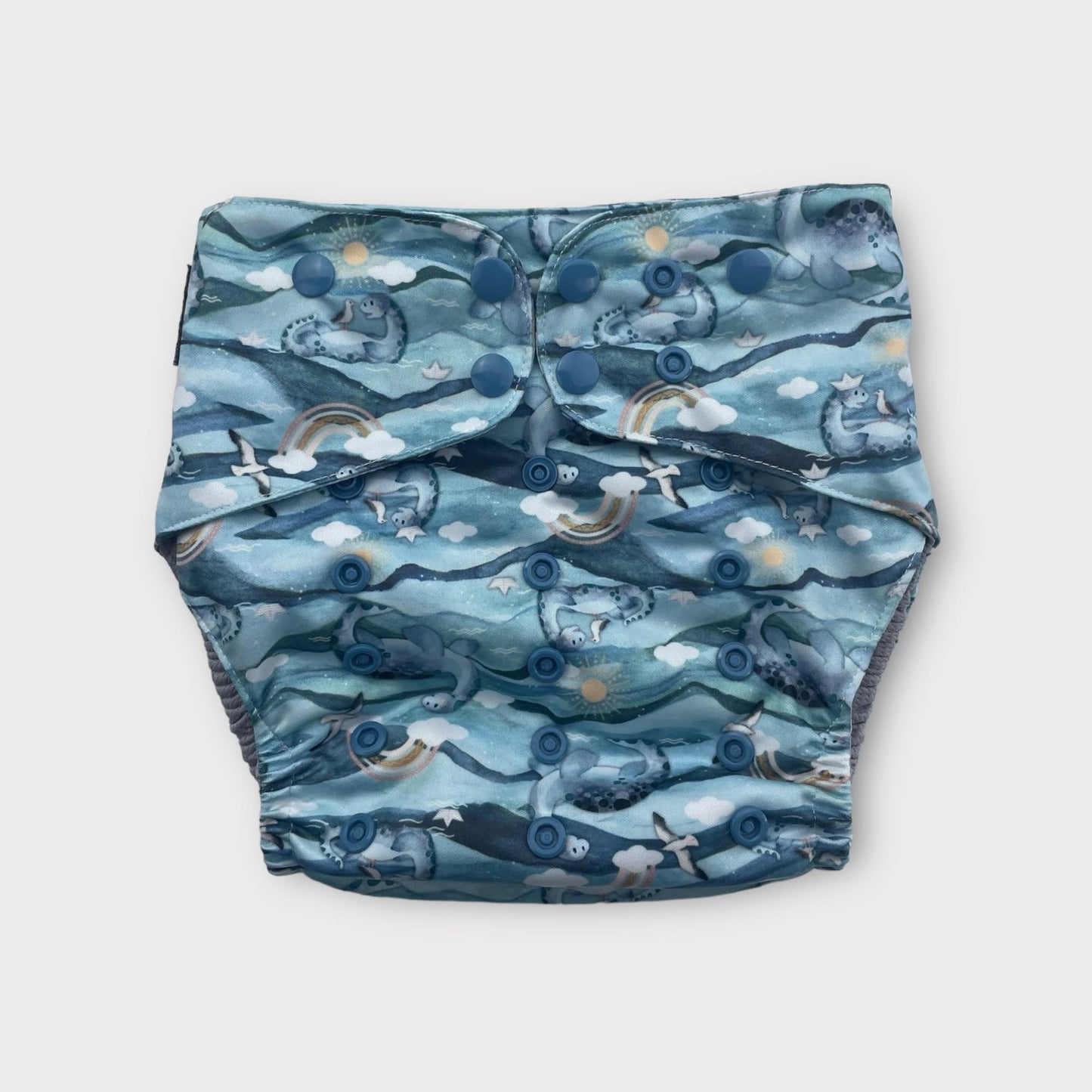 a blue pocket cloth diaper with illustrations of sea gulls, plesiosaurus, brown rainbows, clouds, the sun and water.