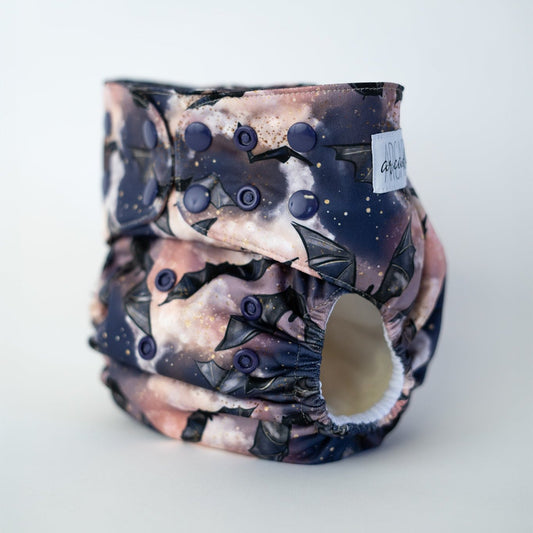 A pocket cloth diaper with a nocturnal theme, featuring a pattern of bats and stars against a cosmic purple background. The snaps are dark blue/purple.