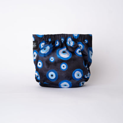 A photo of a cloth diaper standing up with a plain white background. The pattern on the cloth diaper is a light black watercolor, with hand painted Eye illustrations. The eyes are blue, black, and white. This symbol is commonly reffered to as "evil eye" but is known as Mati or Nazar. Rear view.