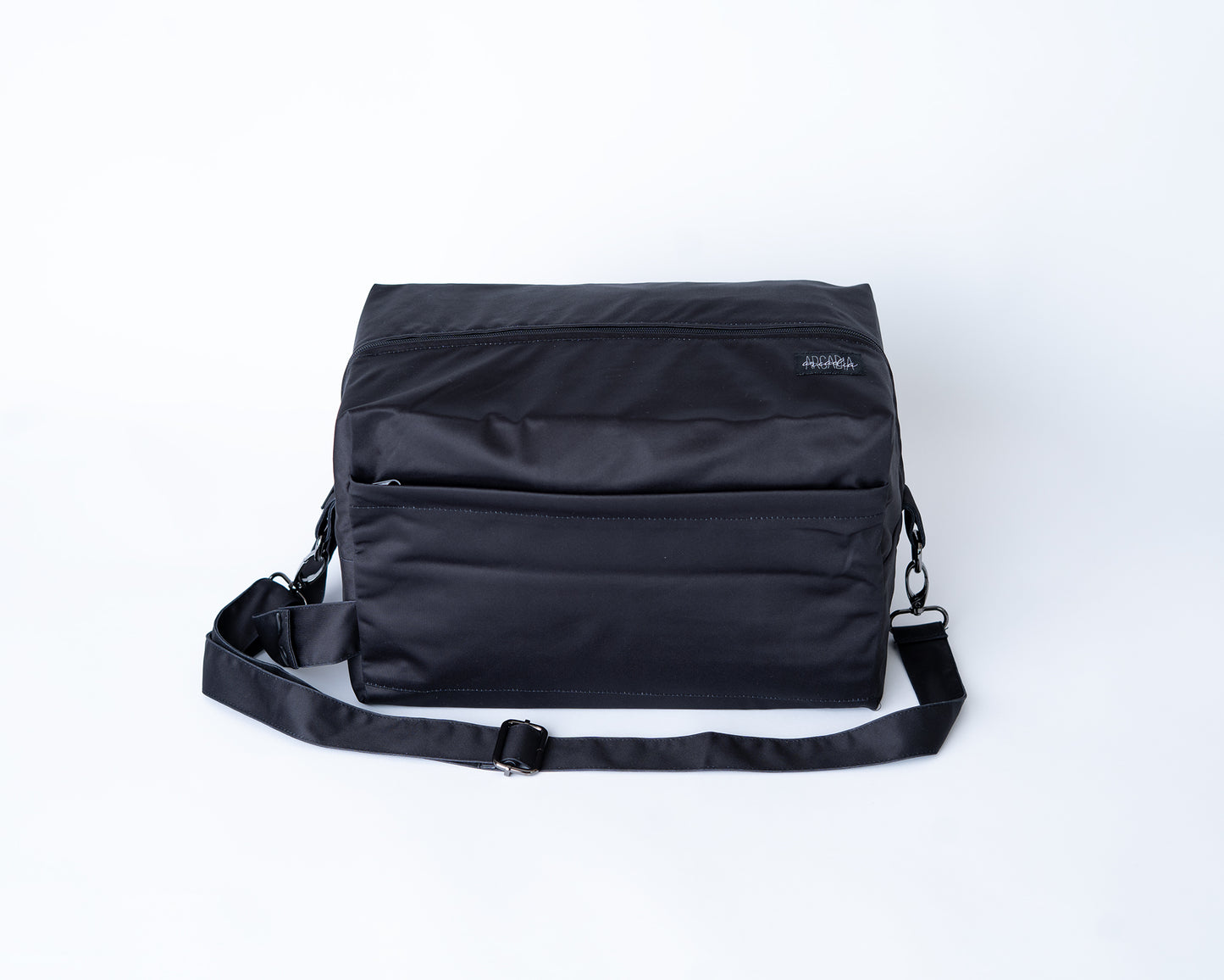Black bag with a strap on a white background. The bag features an adjustable shoulder strap with black metal hardware, a carrying handle, and 2 zipper openings. There is a black Arcadia's nursery logo on the right side of the pod.