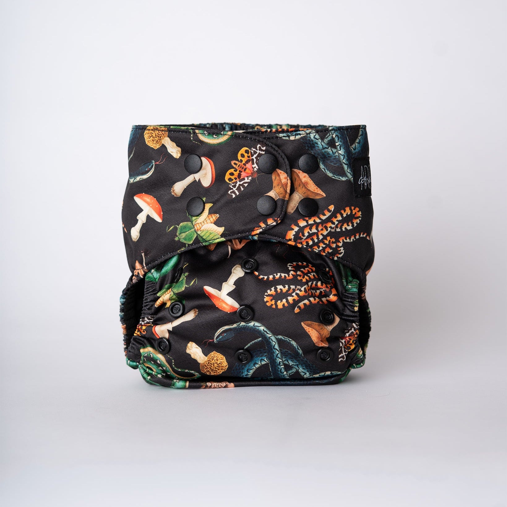 A pocket style cloth diaper is shown straight on against a clean white background. The Ophidian print features coiling snakes, moths, mushrooms, and beetle elements illustrated in rich greens, warm ambers, and earthy neutrals on a dark base. The front panel displays a smooth fit with overlapping matte black snaps visible, highlighting the structured shape and detailed artwork across the center.