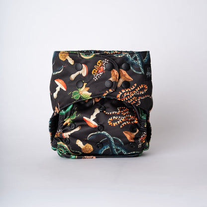 A pocket style cloth diaper is shown straight on against a clean white background. The Ophidian print features coiling snakes, moths, mushrooms, and beetle elements illustrated in rich greens, warm ambers, and earthy neutrals on a dark base. The front panel displays a smooth fit with overlapping matte black snaps visible, highlighting the structured shape and detailed artwork across the center.