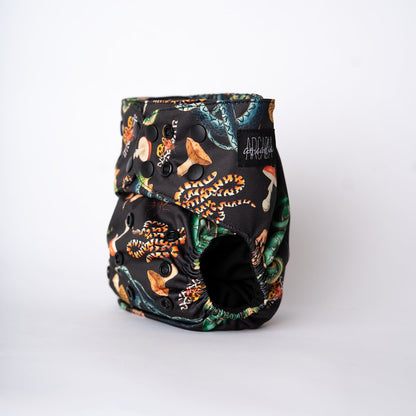 A pocket style cloth diaper is photographed at a slight angle. The print wraps seamlessly around the body, showcasing winding serpents intertwined with mushrooms, leaves, and moths in moody jewel tones and warm earth hues on a dark background. The leg opening and black snaps are visible, emphasizing the contoured fit and craftsmanship of the design as well as a black Arcadia's logo tag.
