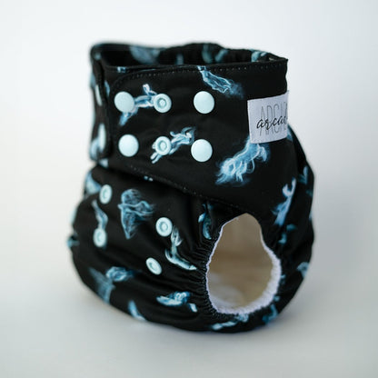 A pocket style cloth diaper in the Patronus print shown at a front angle. The black fabric displays repeating glowing blue spirit animal illustrations. Light blue adjustable snaps are visible along the waist and rise. The leg opening reveals a white AWJ inner lining.
