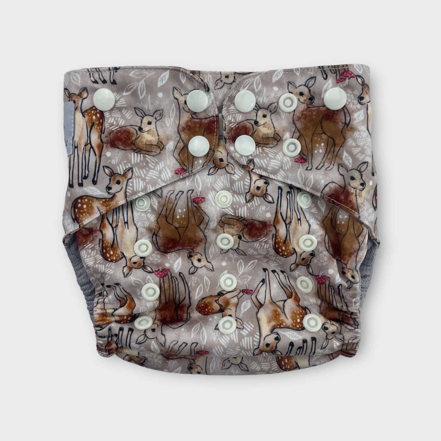 a pocket style cloth diaper with a tan background and drawings of deer with splotches of brown paint. there are also small musrooms and white foliage. the snaps are a light cream color.