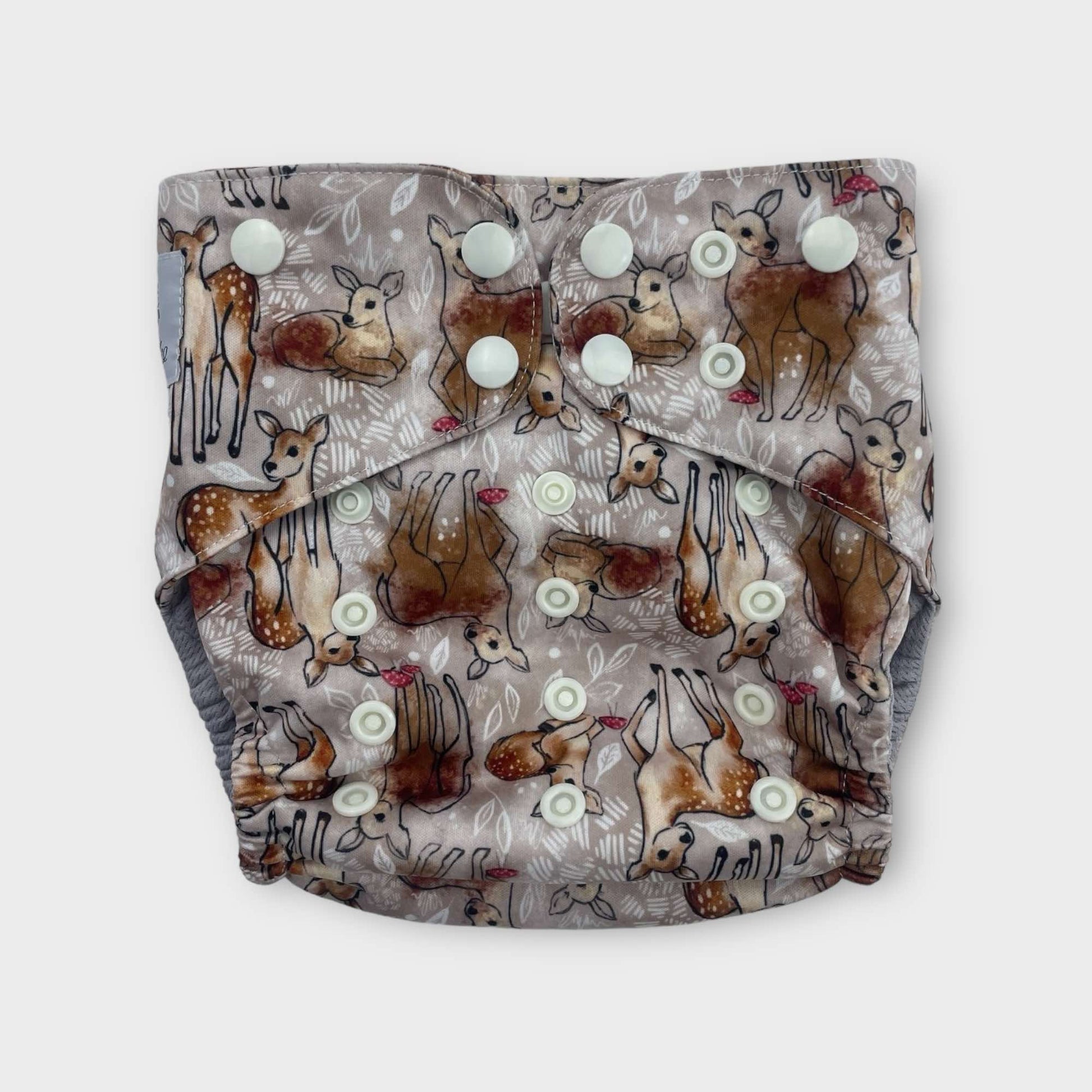 a pocket style cloth diaper with a tan background and drawings of deer with splotches of brown paint. there are also small musrooms and white foliage. the snaps are a light cream color.