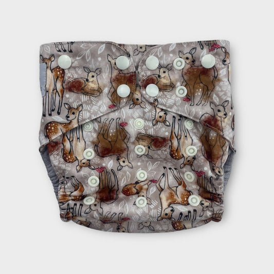 a pocket style cloth diaper with a tan background and drawings of deer with splotches of brown paint. there are also small musrooms and white foliage. the snaps are a light cream color.