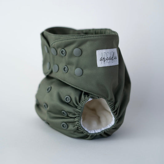 A reusable pocket cloth diaper in a muted, earthy moss green shade named Petrichor. This premium cloth diaper features multiple adjustable snaps for a customizable fit, ensuring comfort and leak protection for babies of all sizes. The soft, elastic leg openings and water-resistant outer layer provide both flexibility and reliability. Front angle view.