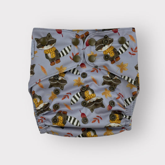 A Wickham Wild cloth diaper with raccoon pattern on a light gray background wearing a scarf and footwear, with fall leaves scattered around the racoon.