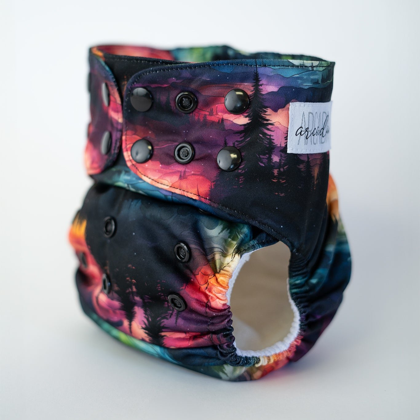 Vibrant cloth diaper showcasing a stunning landscape design. The rich blend of colors, from deep purples and blues to fiery reds and oranges, creates a mesmerizing aurora effect over a forested mountain scene. This reusable diaper offers both artistic flair and comfort for your baby. Front angle view.