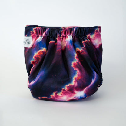 The back view of a pocket style cloth diaper in the Starburst print, photographed against a clean white background. The design features a bold cosmic explosion of deep navy, electric magenta, and glowing white streaks that radiate like a celestial flare across the fabric. The gathered elastic waistband creates a secure fit while highlighting the dramatic, galaxy inspired artwork.