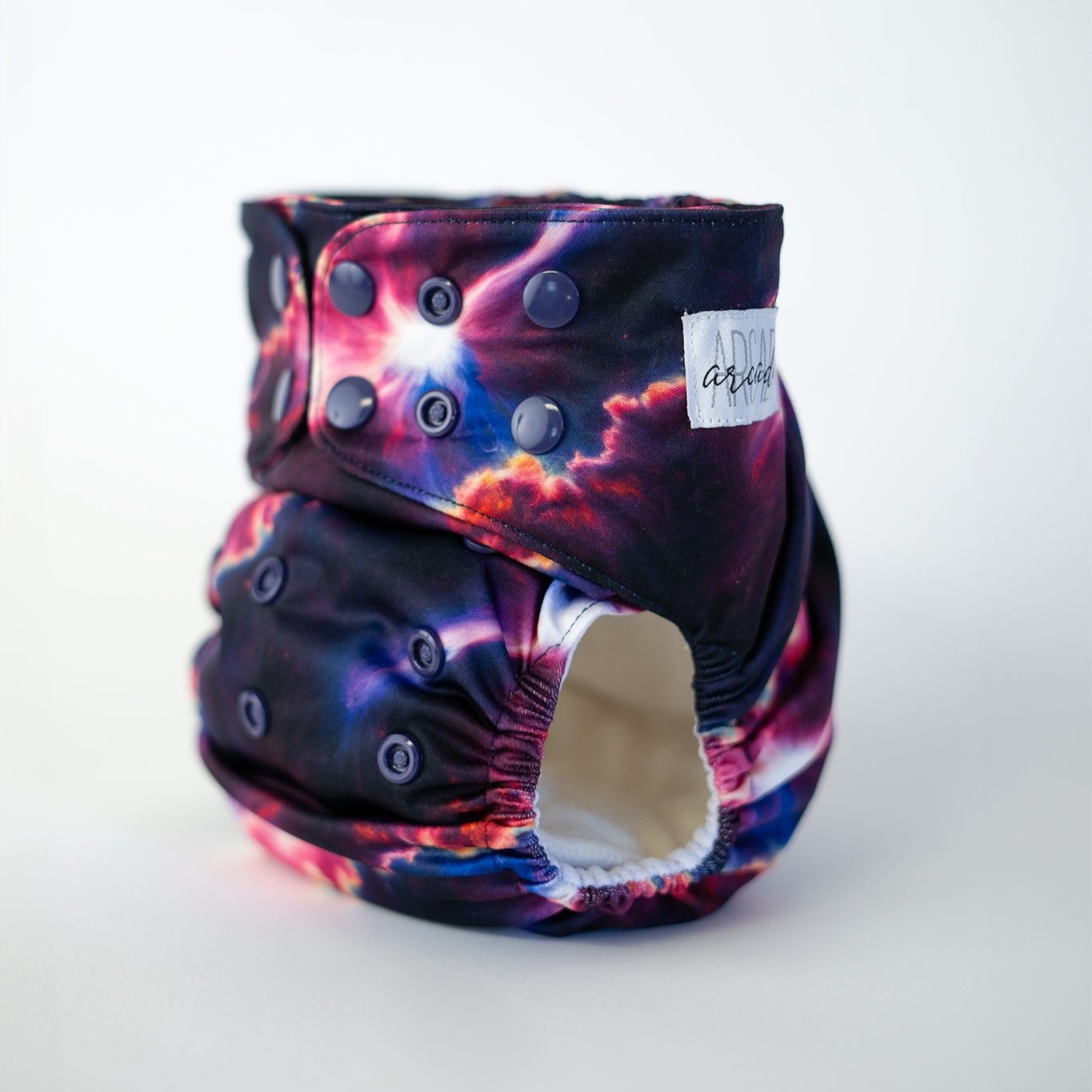 A pocket cloth diaper in the Starburst print shown at a front angle, revealing adjustable snap closures along the waist and rise. The print showcases a striking cosmic burst pattern with rich blues, vivid pinks, and bright star like highlights that blend into a dark night sky backdrop. Contoured leg elastics and structured shaping emphasize both performance and an otherworldly aesthetic.