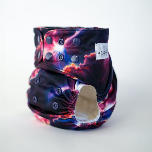 A pocket cloth diaper in the Starburst print shown at a front angle, revealing adjustable snap closures along the waist and rise. The print showcases a striking cosmic burst pattern with rich blues, vivid pinks, and bright star like highlights that blend into a dark night sky backdrop. Contoured leg elastics and structured shaping emphasize both performance and an otherworldly aesthetic.
