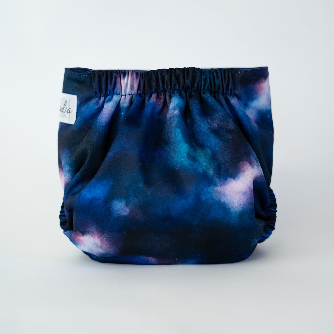 The back view of an Arcadia’s Nursery pocket cloth diaper in the Stardust print, displayed against a white background. The fabric features a cosmic design with deep navy and cerulean blue tones blended with cloudy pink. Gathered elastic at the waist and legs creates a secure, contoured fit while showcasing the galaxy inspired artwork.