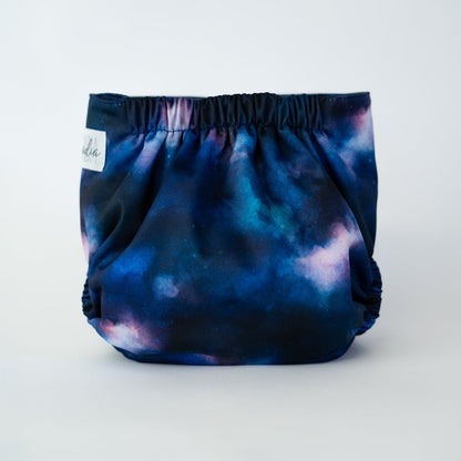 The back view of an Arcadia’s Nursery pocket cloth diaper in the Stardust print, displayed against a white background. The fabric features a cosmic design with deep navy and cerulean blue tones blended with cloudy pink. Gathered elastic at the waist and legs creates a secure, contoured fit while showcasing the galaxy inspired artwork.