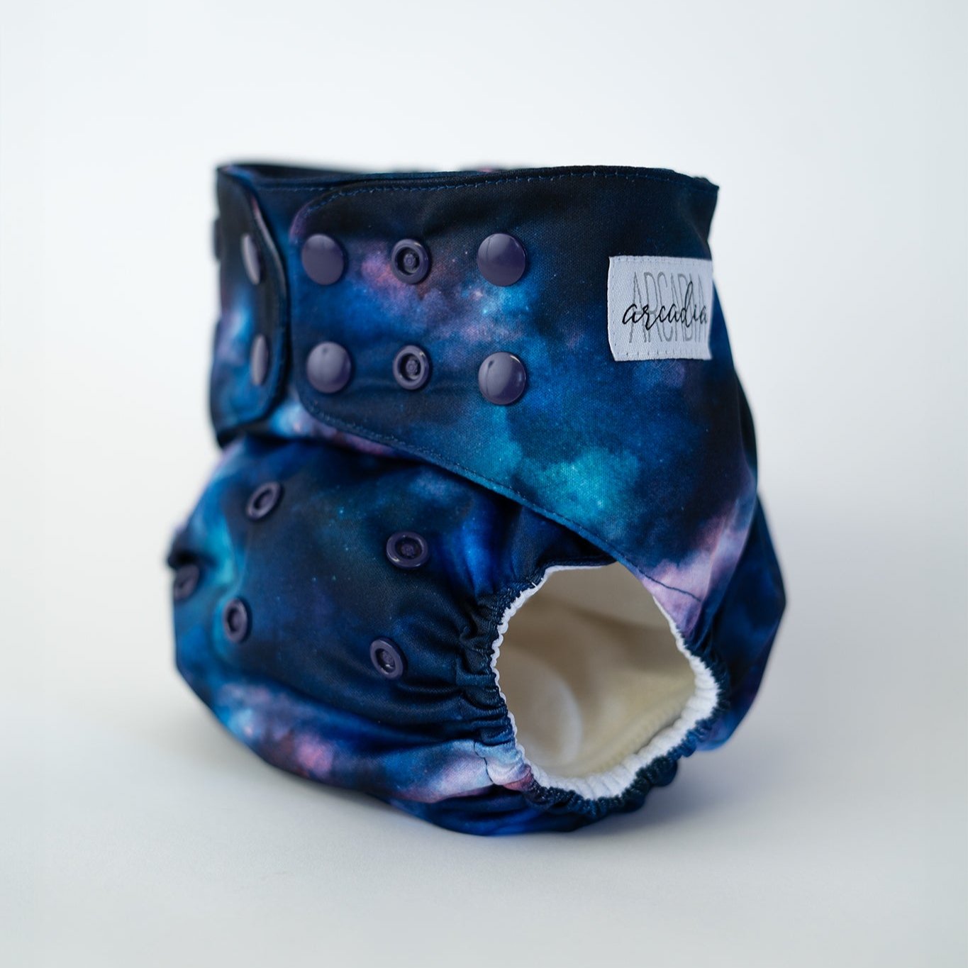 A pocket style cloth diaper from Arcadia’s Nursery shown at a slight front angle in the Stardust print. The cosmic fabric displays rich blues, inky shadows, and cloudy pink blotches for a galactic look. Coordinating blue snap closures along the waist and rise provide an adjustable fit, and the curved leg openings highlight the sculpted, performance focused design