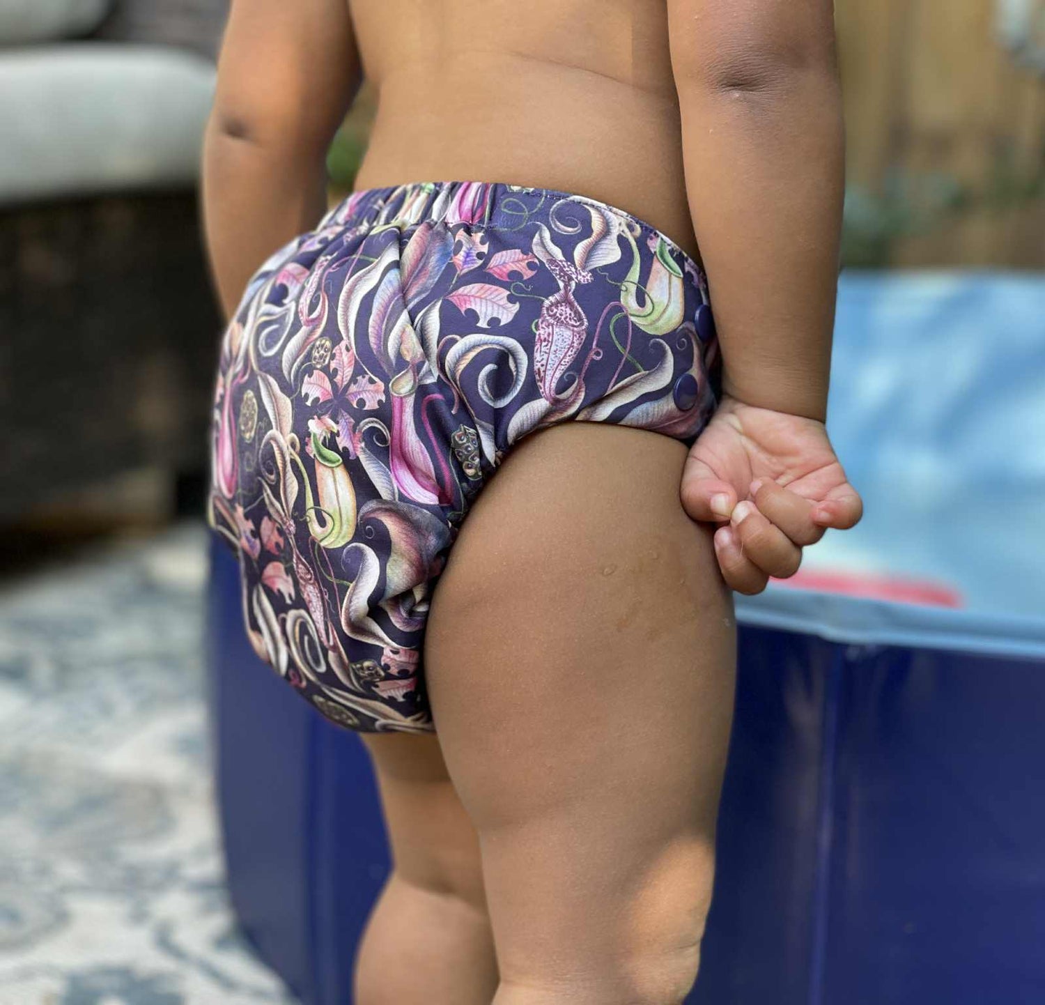 Toddler wearing a cloth diaper standing near a blue pool outdoors. The cloth diaper is the print Trapped, it features illustrations of carnivorous plants and pitcher plants with a deep purpleish blue background.