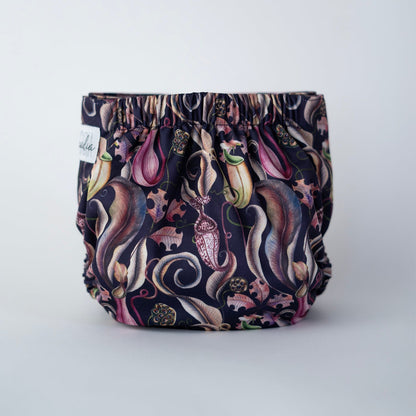 A standing cloth diaper from the back view is displayed against a clean white background. The fabric showcases a striking print of pitcher plants, leaves, and seed pods on a  purple/blue base. Coordinating deep purple-blue snaps complement the bold, carnivorous plant design. The gentle back elastic is visible