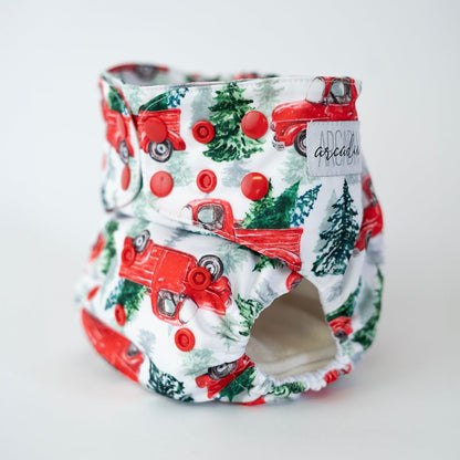 An angled front view of an Arcadia’s Nursery pocket style cloth diaper featuring a winter print. Red snap closures line the waist and rise for an adjustable fit. The background of the print is white, the print features illustrations of vintage red trucks with Christmas trees in the bed of the truck. The curved leg opening with soft elastic and the Arcadia’s Nursery woven label are visible, highlighting both the functional construction and the crisp, seasonal aesthetic.