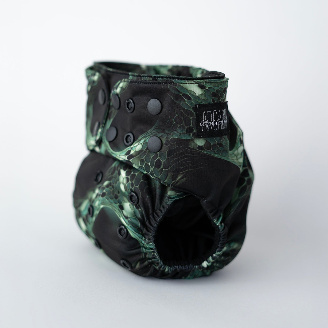 Reptile inspired pocket cloth diaper featuring a sleek green and black scale like pattern, resembling interconnected cells on a black background. This reusable diaper features black snap closure, a black logo tag, and comfortable elastics. Front angle view.