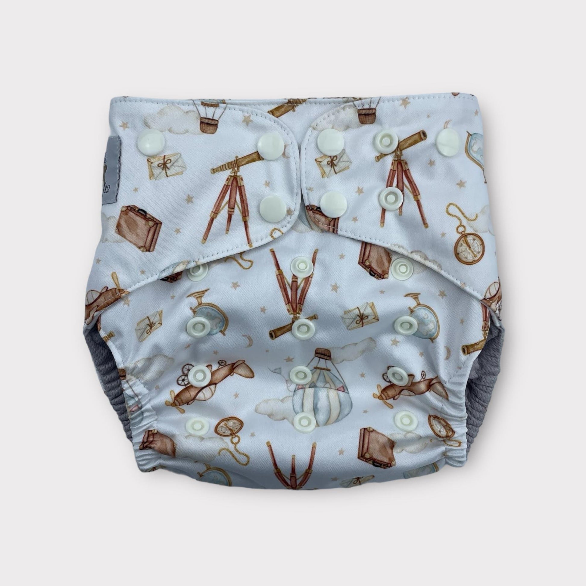 A pocket style wickham wild reusable diaper with a light blue background. There are illustrations of antique items like a pocketwatch, a globe, suitase, telescope, hot air balloon, Letters, and an old airplane.