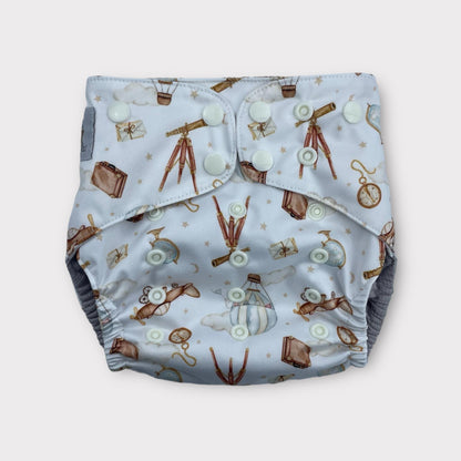 A pocket style wickham wild reusable diaper with a light blue background. There are illustrations of antique items like a pocketwatch, a globe, suitase, telescope, hot air balloon, Letters, and an old airplane.