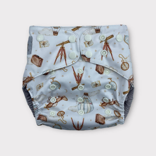 A pocket style wickham wild reusable diaper with a light blue background. There are illustrations of antique items like a pocketwatch, a globe, suitase, telescope, hot air balloon, Letters, and an old airplane.