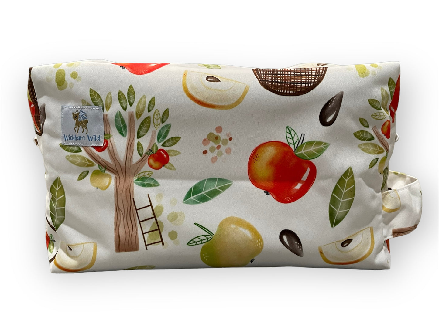 a diaper pod bag with a light bakground and apple orchard illustrations. This print features an apple tree with a ladder resting against it, whole apples, sliced apples, and seeds. A wickham wild logo tag is visible in the photo which is sewn onto the pod.