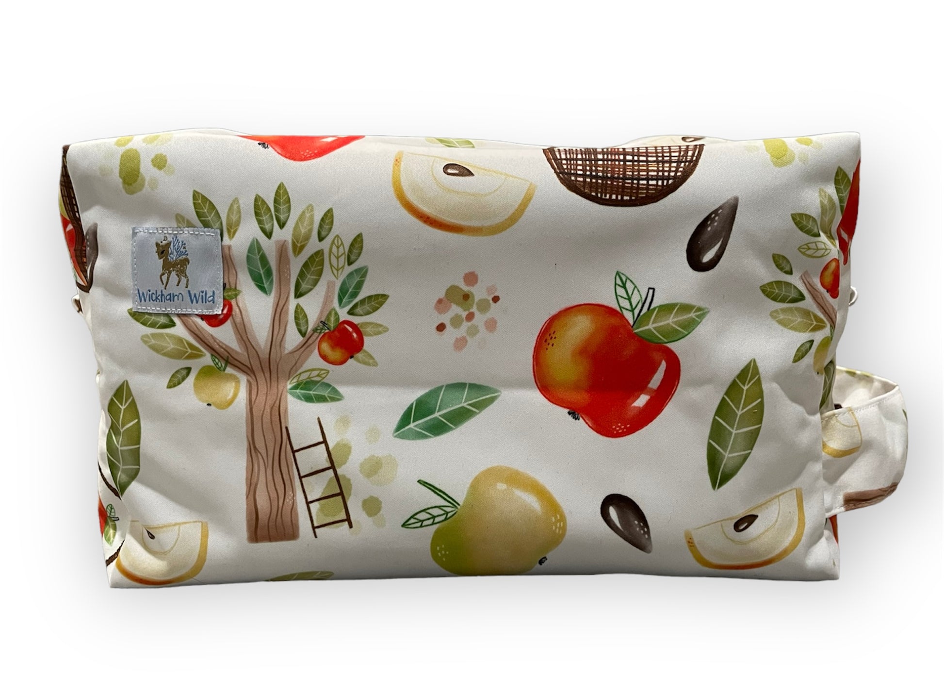 a diaper pod bag with a light bakground and apple orchard illustrations. This print features an apple tree with a ladder resting against it, whole apples, sliced apples, and seeds. A wickham wild logo tag is visible in the photo which is sewn onto the pod.