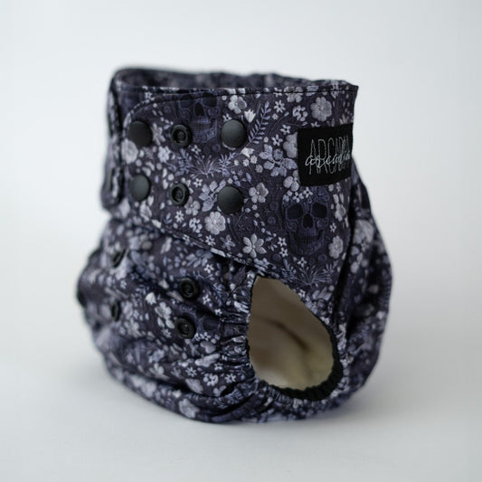 A black pocket diaper with a floral pattern, featuring adjustable snap buttons and a brand tag.
