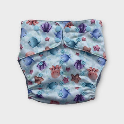 A pocket style cloth diaper. The print background is white with light blue bubbles. The illustrations on the diapers are cute cartoony watercolor ocean critters like sea stars, clams, octopus, and fish. the snaps are light blue.