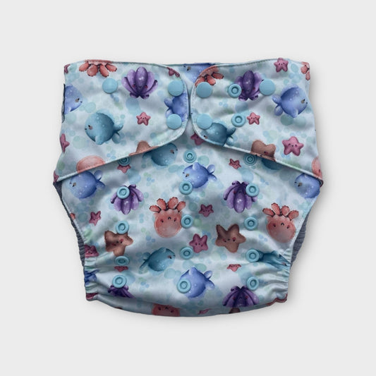 A pocket style cloth diaper. The print background is white with light blue bubbles. The illustrations on the diapers are cute cartoony watercolor ocean critters like sea stars, clams, octopus, and fish. the snaps are light blue.