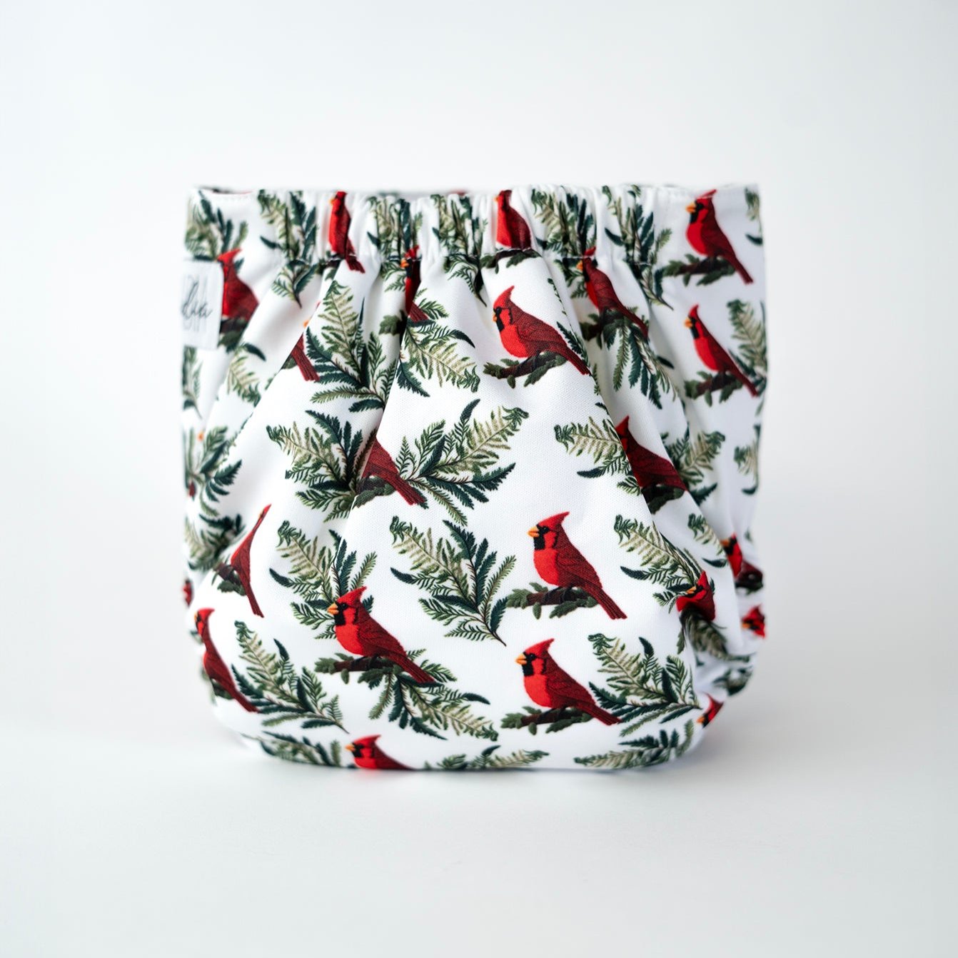The back view of an Arcadia’s Nursery pocket style cloth diaper shown against a clean white background. The print features a white background, With images of red cardinals perched on branches of pine trees. Gathered elastic along the waist and legs creates a gently contoured shape, while the fabric displays the wintery cardinal/bird pattern uninterrupted.