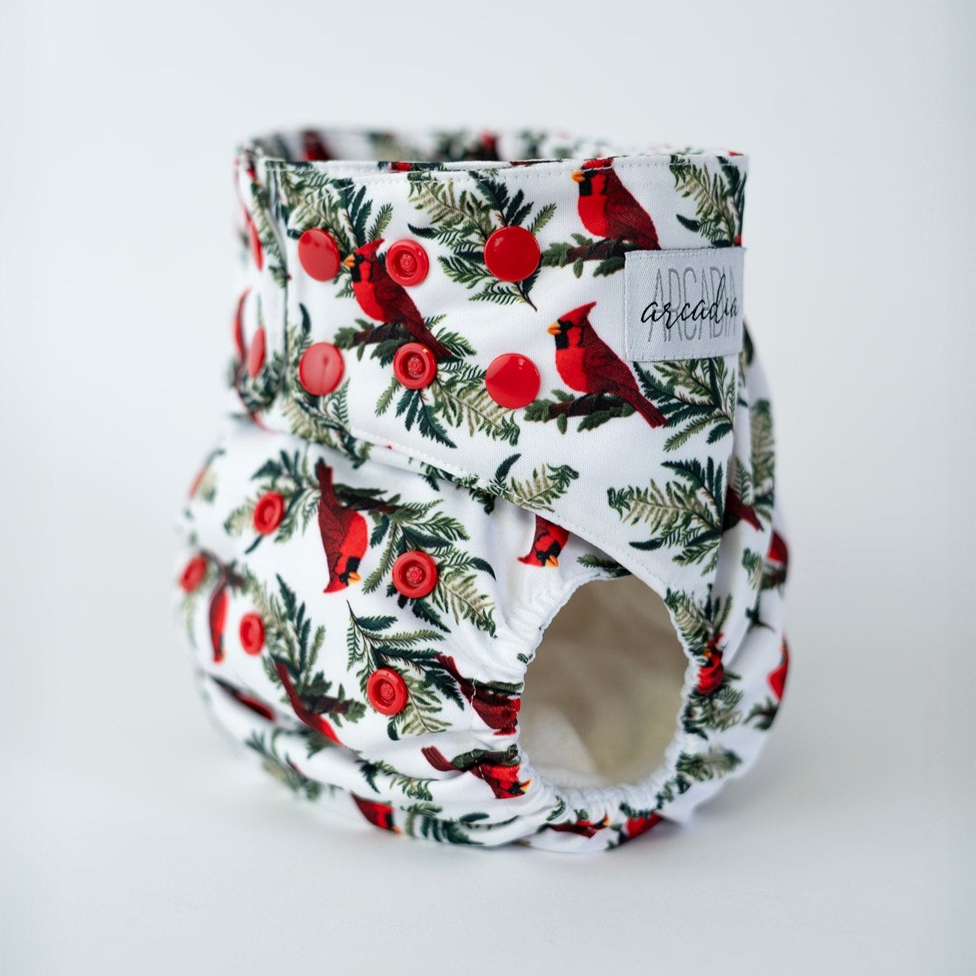 An angled front view of a pocket style cloth diaper featuring winter-ish cardinal bird print. Red snap closures line the waist and rise for an adjustable fit. The print has a solid white background with illustrations of red cardinals on branches of coniferous/pine trees. The curved leg opening with elastic and the Arcadia’s Nursery logo tag are visible, highlighting both the functional construction.