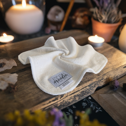 This styled product photo features a reusable cloth wipe laid on a rustic wooden surface. The wipe is white with a soft, plush texture. A fabric label is sewn into one corner, displaying the brand name Arcadia, and noting the material composition: "85% Bamboo, 15% Polyester." The scene is warmly lit, with glowing candles in the background, creating a cozy, inviting atmosphere. Surrounding elements, such as dried flowers and pottery.