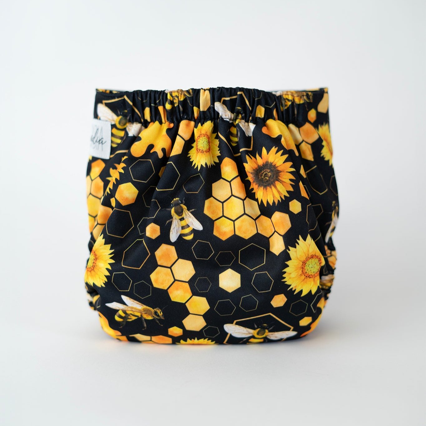 The back view of an Arcadia’s Nursery pocket style cloth diaper displayed against a white background. The print features golden honeycomb hexagons, sunflowers, and flying bees layered over a deep black base. Warm yellow and amber tones stand out vividly, with gathered elastic along the waist and legs creating a softly contoured fit designed for comfort and coverage.