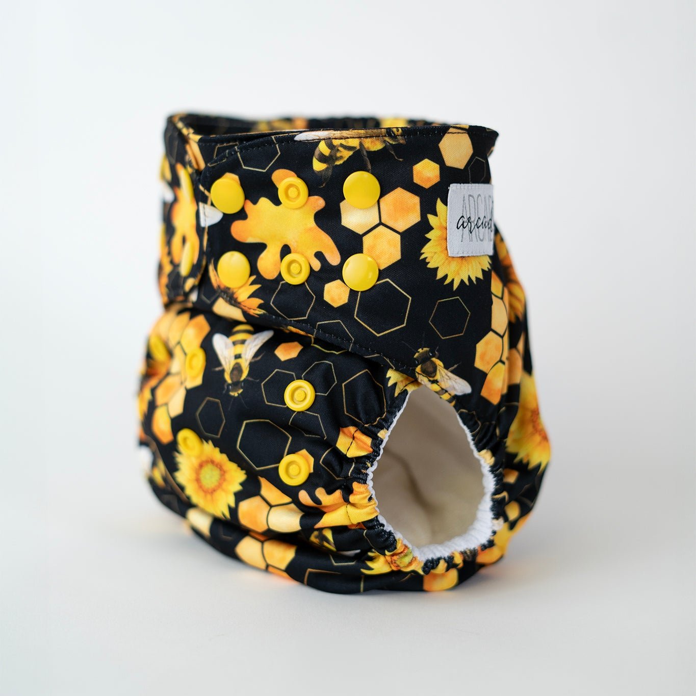 An angled front view of an Arcadia’s Nursery pocket style cloth diaper, highlighting the snap closures and leg openings. Bright yellow snaps run along the waist and rise for adjustability, while the honeybee and sunflower print continues seamlessly across the front panel. The curved shape and snug leg elastics are visible, emphasizing both the functional design and the bold, nature inspired pattern.
