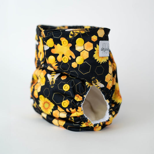 An angled front view of an Arcadia’s Nursery pocket style cloth diaper, highlighting the snap closures and leg openings. Bright yellow snaps run along the waist and rise for adjustability, while the honeybee and sunflower print continues seamlessly across the front panel. The curved shape and snug leg elastics are visible, emphasizing both the functional design and the bold, nature inspired pattern.